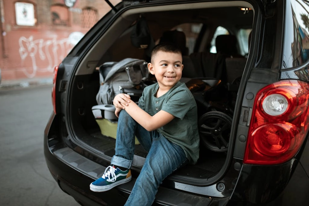 Smiling child sitting in the trunk of an SUV on an urban street, wearing casual clothes.
